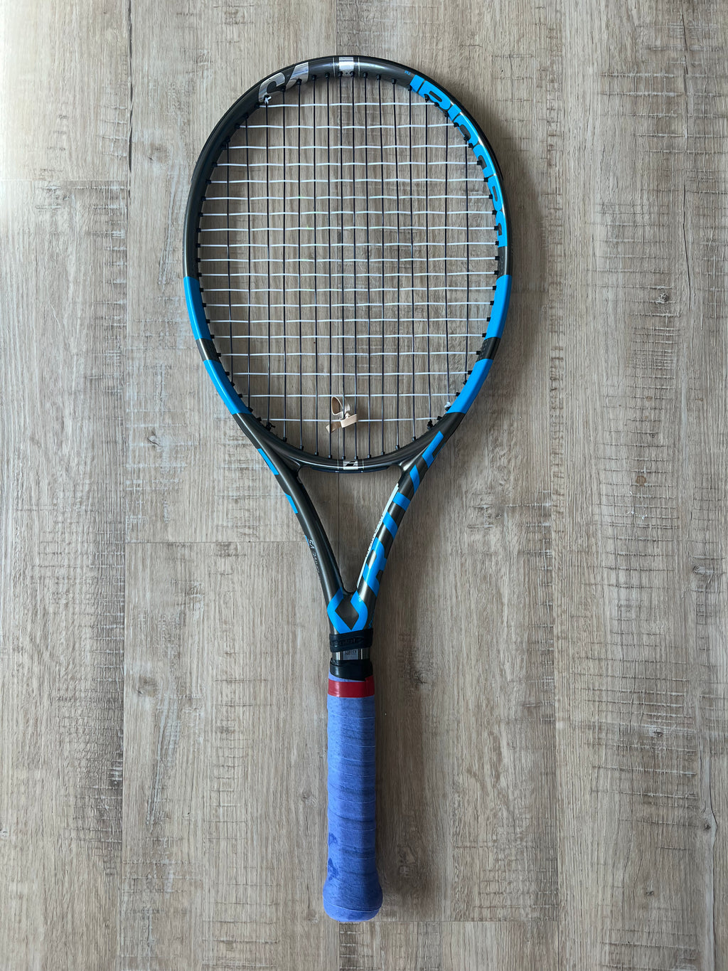 A pre-owned Babolat Pure Drive VS 4 3/8 tennis racket with blue grip tape rests on a wooden floor—ideal for advanced players seeking precision and power.