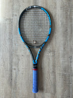 A pre-owned Babolat Pure Drive VS 4 3/8 tennis racket with blue grip tape rests on a wooden floor—ideal for advanced players seeking precision and power.