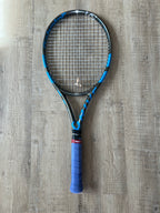 A pre-owned Babolat Pure Drive VS tennis racket (4 3/8 grip) lies on a wooden floor, ideal for advanced players.
