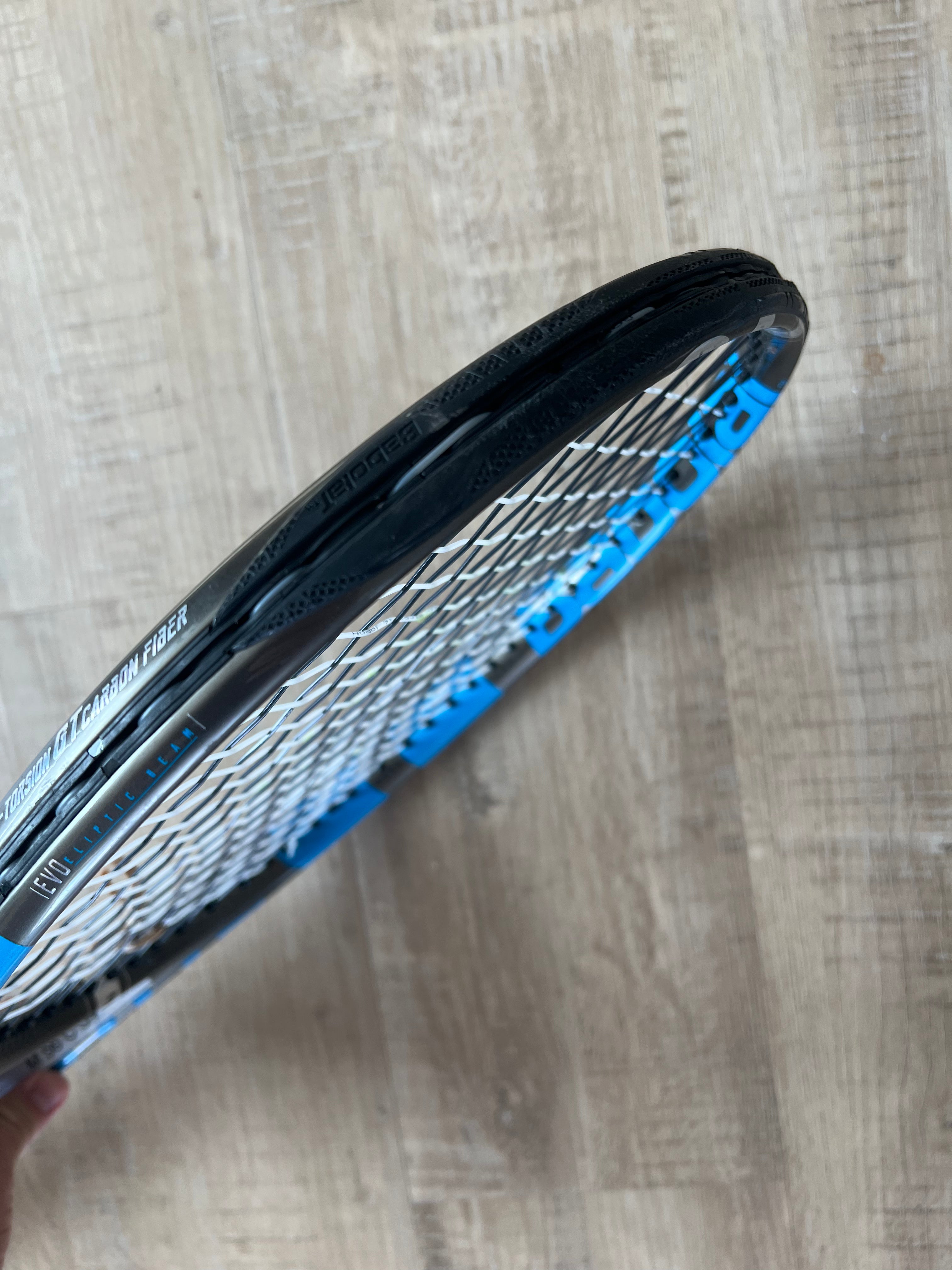 A close-up of a pre-owned Babolat Pure Drive VS (4 3/8) tennis racket with a bent frame on a light wood floor—perfect for advanced players seeking quality Babolat gear.