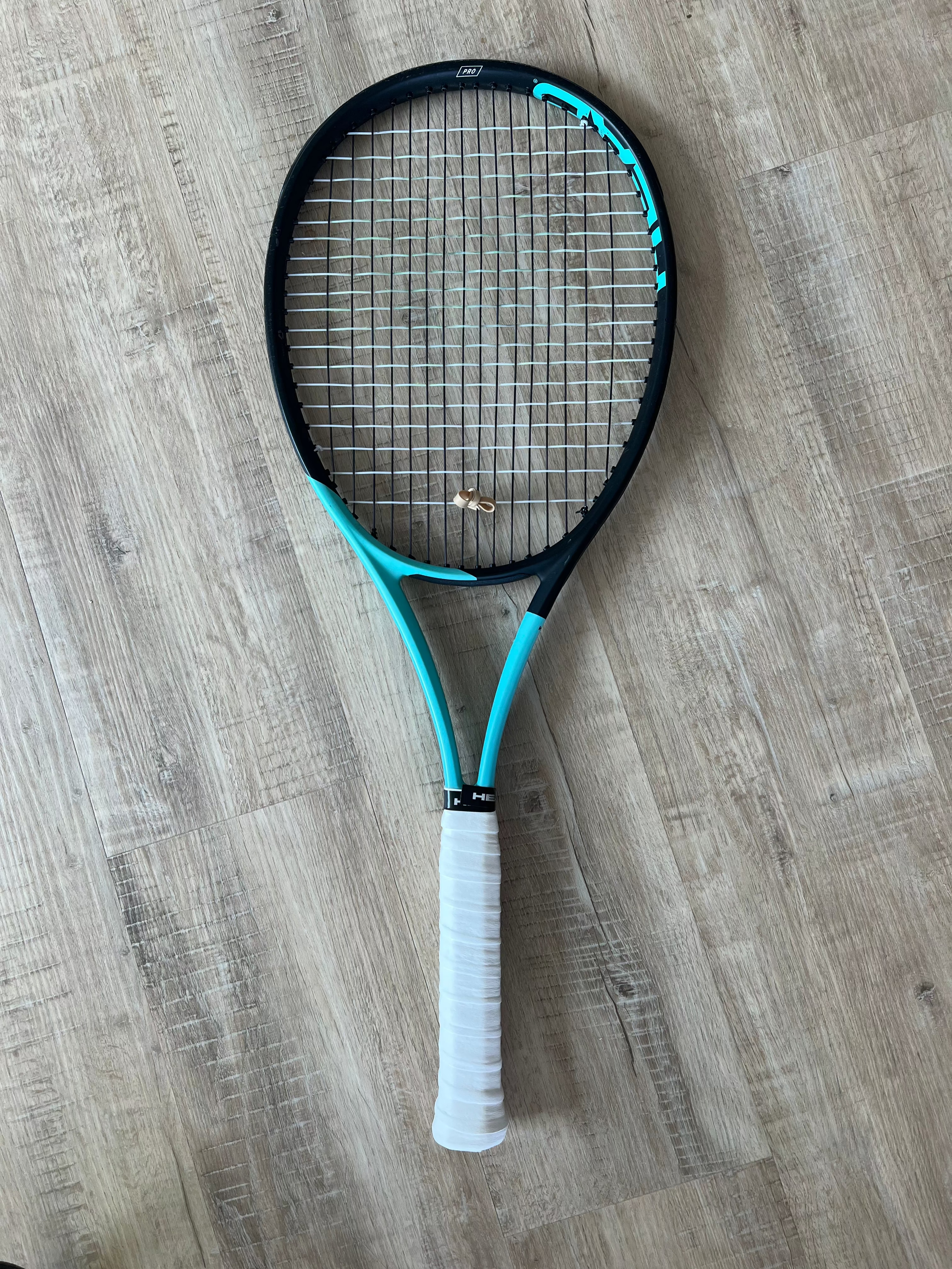 A pre-owned Head Boom Pro 2022 (4 3/8) tennis racquet with a blue and black frame and white grip handle is on a light wooden floor, ideal for advanced players. Two small rings rest on its strings.