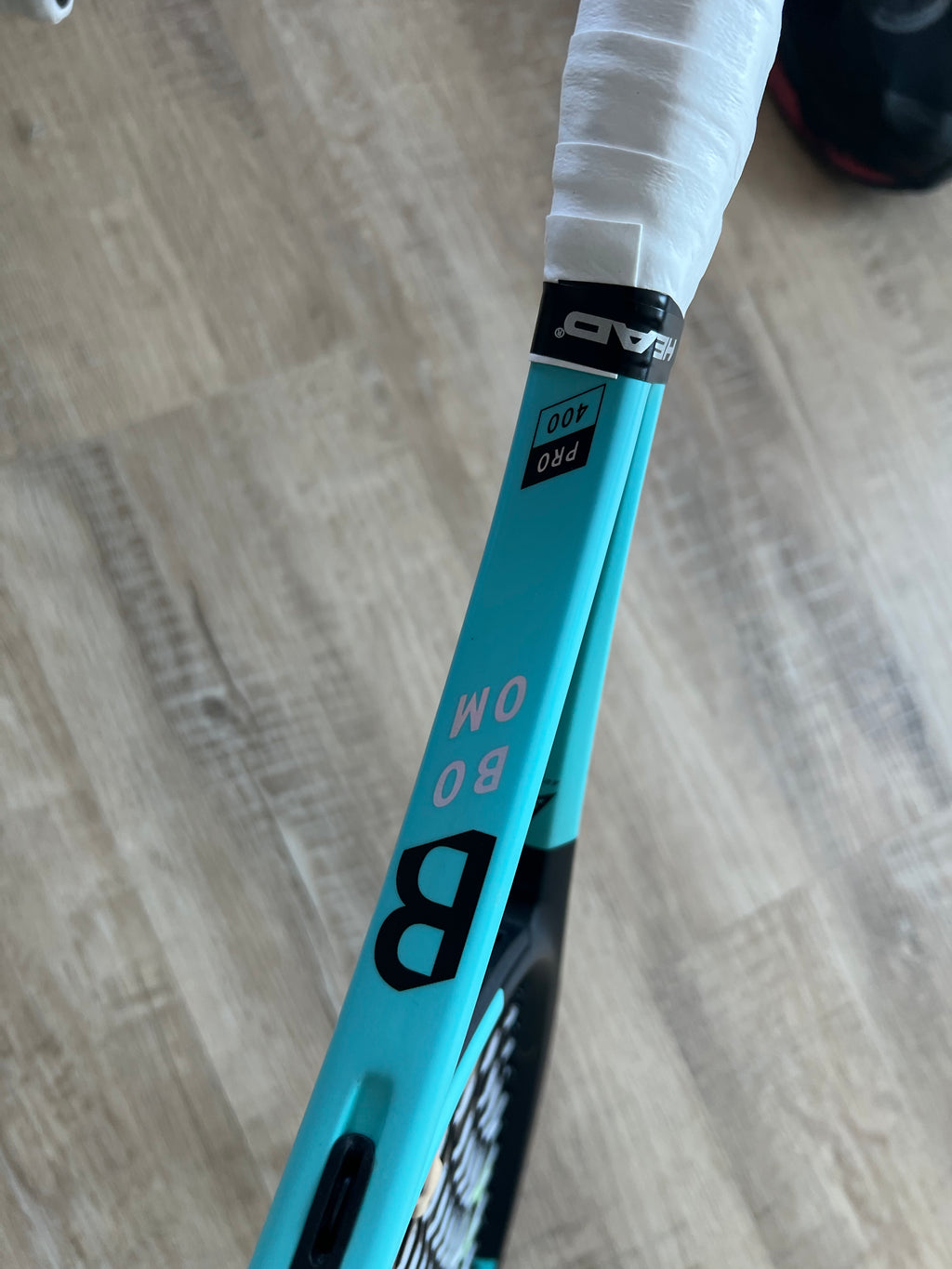 A close-up of a turquoise Head Boom Pro 2022 (Pre-Owned) 4 3/8 tennis racquet with "PRO 100," "BOM," and "B" on the frame, set on a light wooden floor—perfect for advanced players.