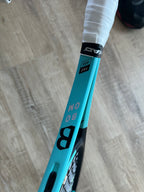 A close-up of a turquoise Head Boom Pro 2022 (Pre-Owned) 4 3/8 tennis racquet with "PRO 100," "BOM," and "B" on the frame, set on a light wooden floor—perfect for advanced players.