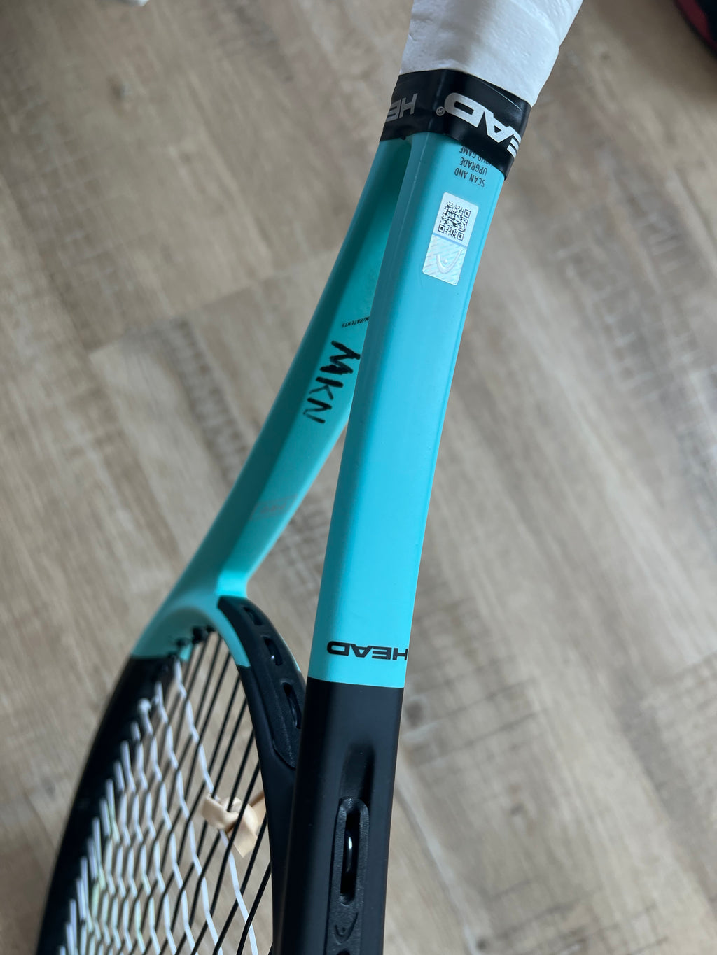 A close-up of a turquoise Head Boom Pro 2022 (Pre-Owned) 4 3/8 tennis racquet shows the brand name, QR code sticker, and part of the handle on a wooden floor—great for advanced players seeking high performance.