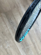 Close-up of a Head Boom Pro 2022 (Pre-Owned) 4 3/8 tennis racquet with a loose bumper guard and visible strings, set on a wooden floor—great for advanced players attentive to racquet wear.