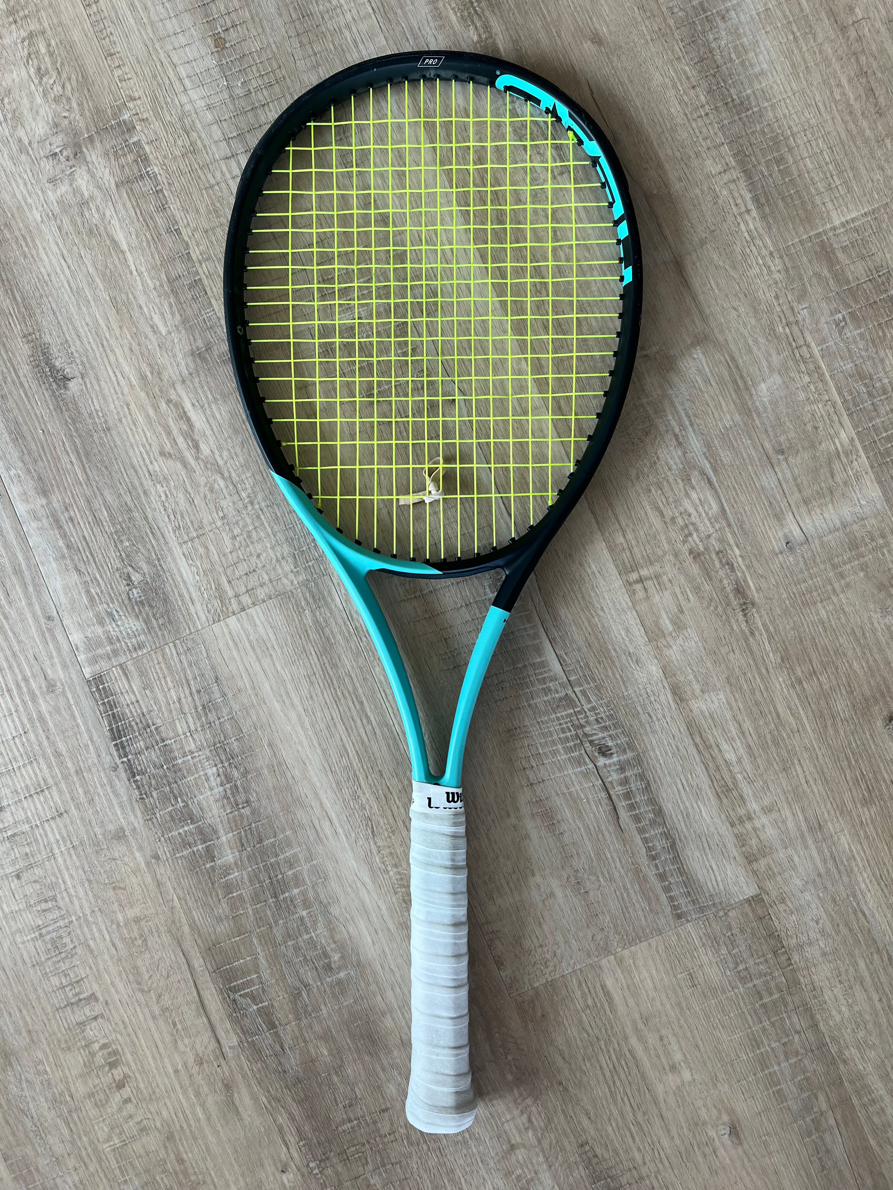 A pre-owned Head Boom Pro 2022 (4 1/4) tennis racquet, featuring a black and teal frame with yellow strings and a white grip, lies on a wooden floor—ideal for aggressive players who want power and precision.
