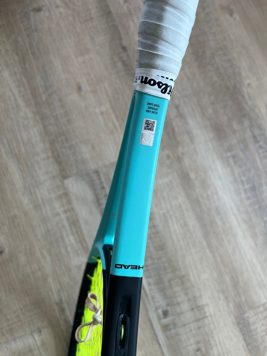 A close-up of a pre-owned Head Boom Pro 2022 (4 1/4) tennis racquet handle with white grip tape and "HEAD" branding, featuring a barcode sticker, rests on a wooden floor—designed for aggressive players.