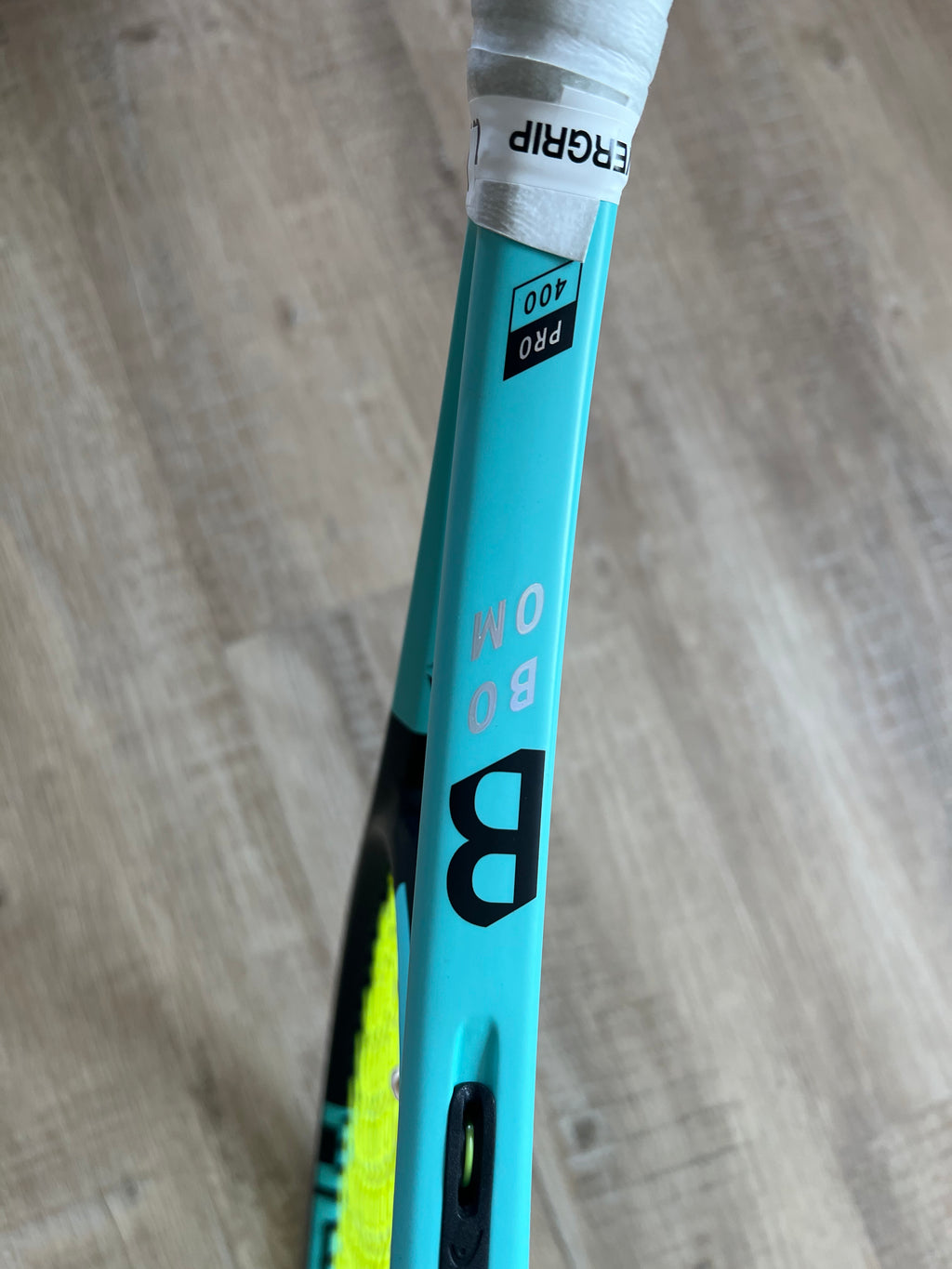 A close-up of a pre-owned Head Boom Pro 2022 (4 1/4) tennis racquet with “B OOM” and “PRO 400” on the frame, resting on a light wooden floor—ideal for aggressive players who want power and precision.
