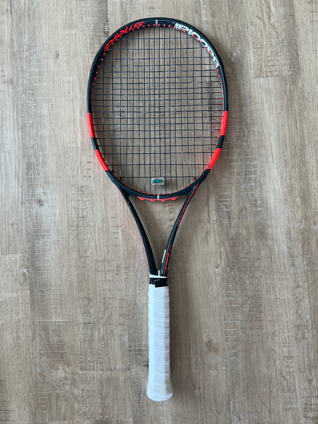 A black and red tennis racket with a white grip is placed on a wooden floor.