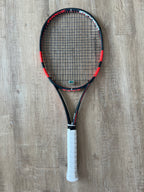 A black and red tennis racket with a white grip is placed on a wooden floor.