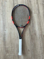 A black and red tennis racket with a white grip is placed on a wooden floor.