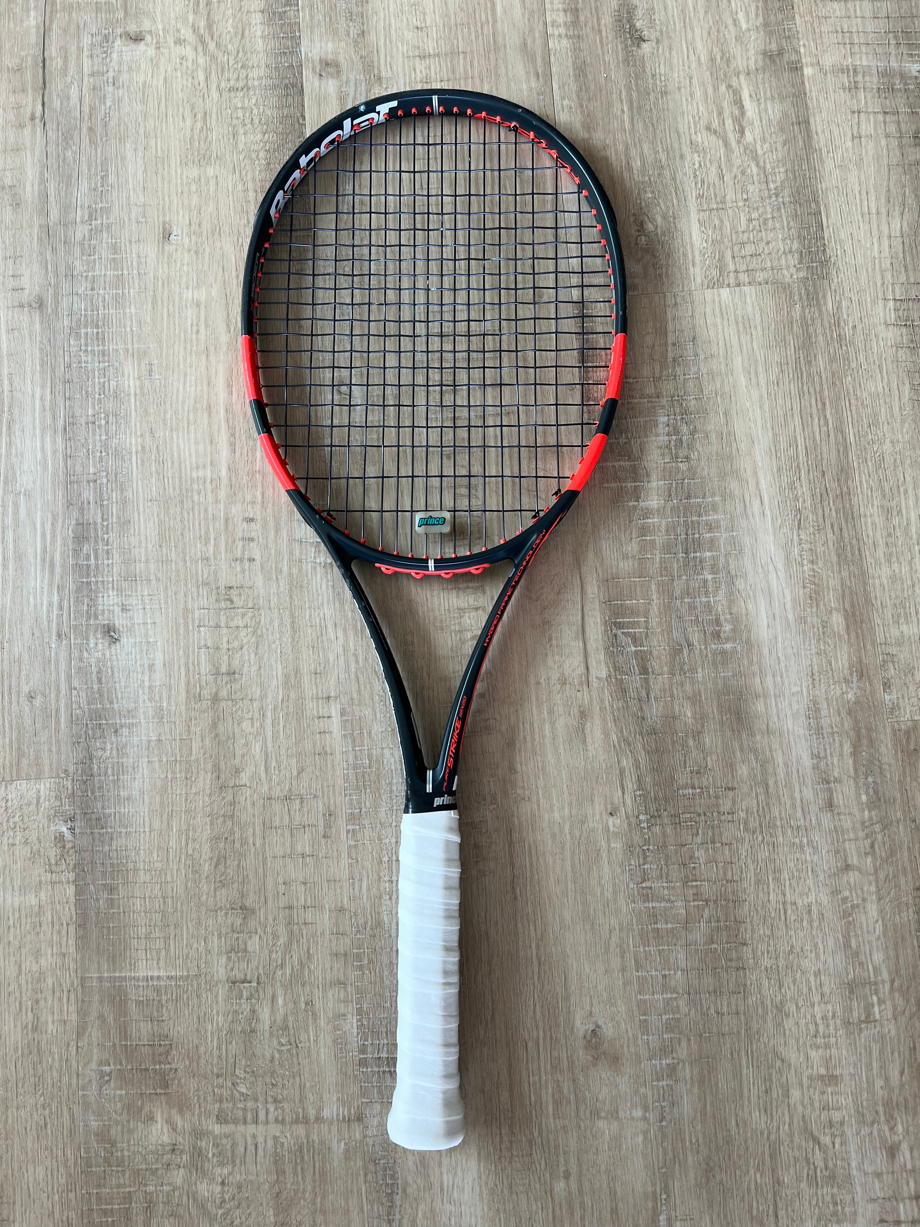 A black and red tennis racket with a white grip is placed on a wooden floor.