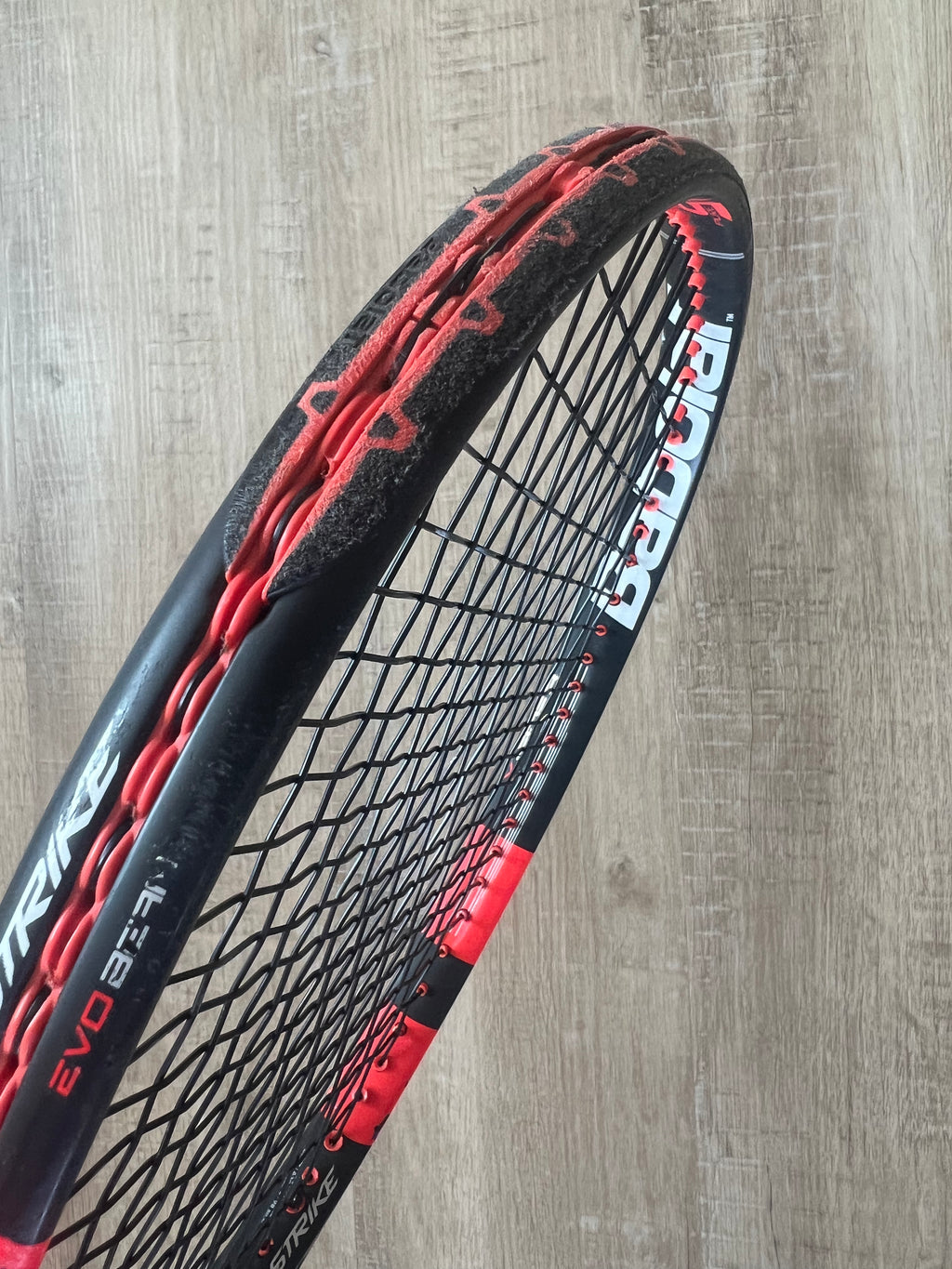 A close-up of a black and red tennis racquet with frayed and worn strings against a wooden background.