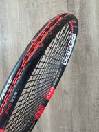 A close-up of a black and red tennis racquet with frayed and worn strings against a wooden background.