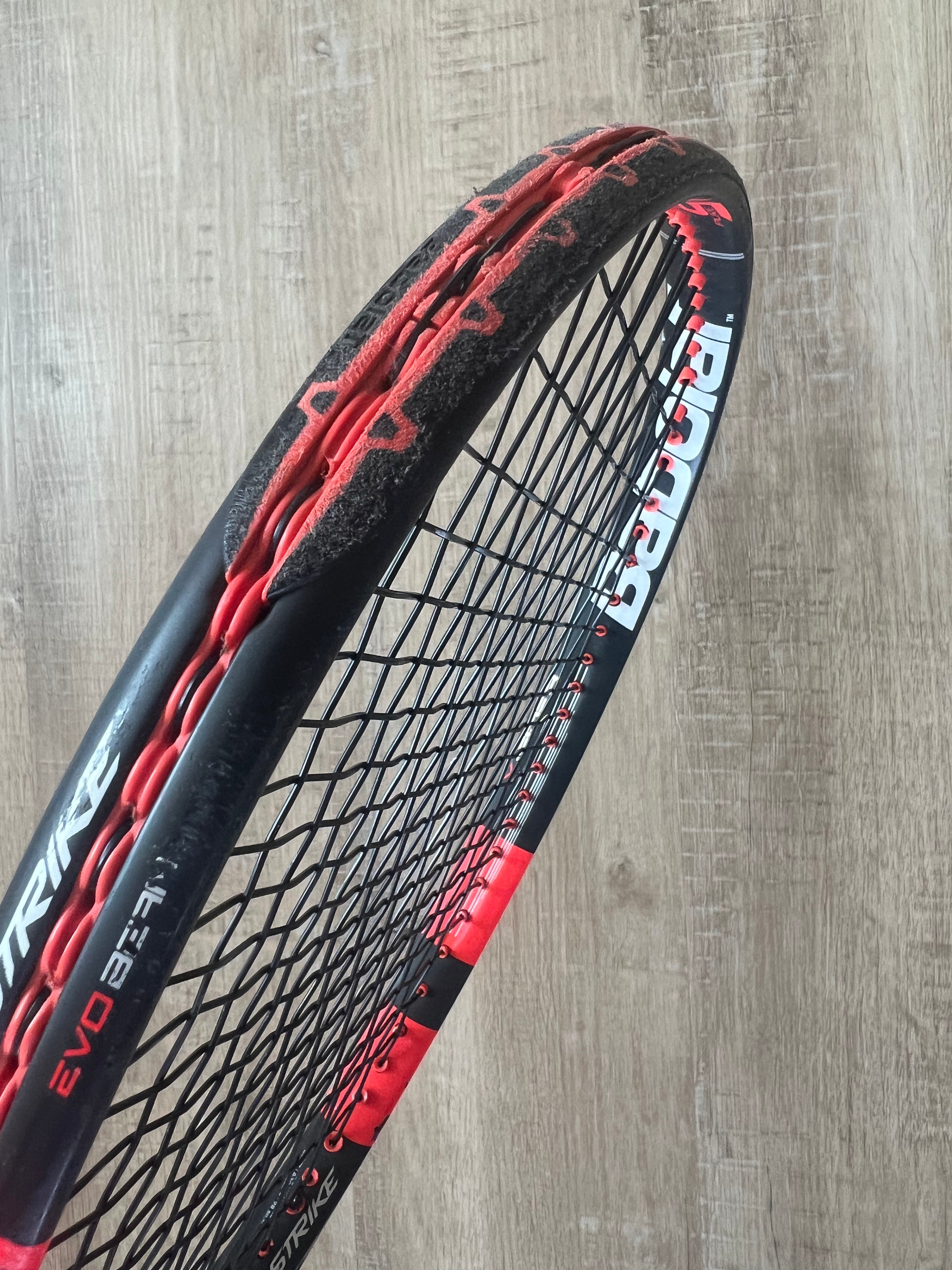 A close-up of a black and red tennis racquet with frayed and worn strings against a wooden background.
