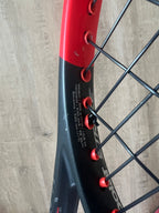 Close-up of a black and red tennis racket, showing string pattern and specifications printed on the frame against a wooden background.