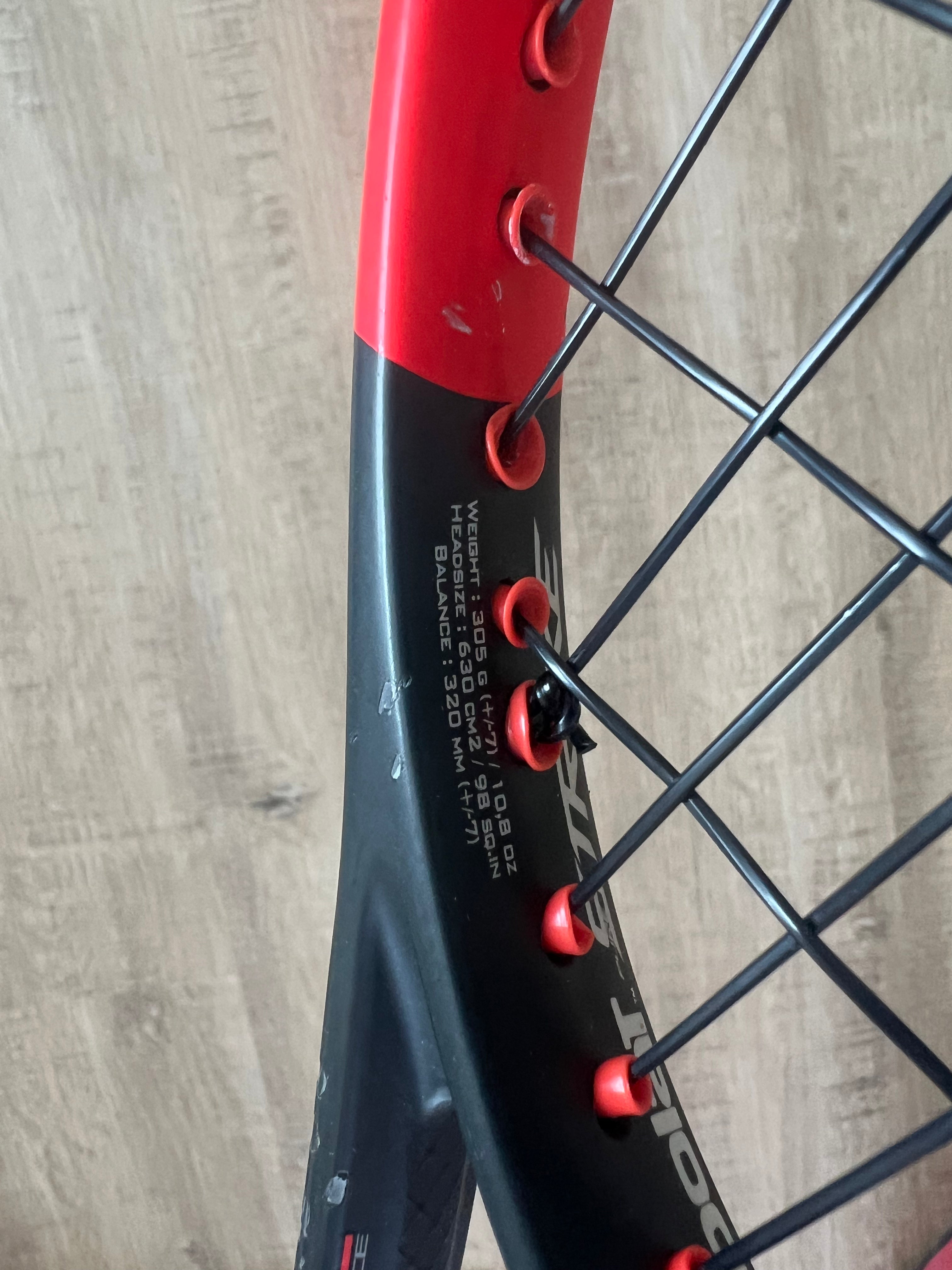 Close-up of a black and red tennis racket, showing string pattern and specifications printed on the frame against a wooden background.
