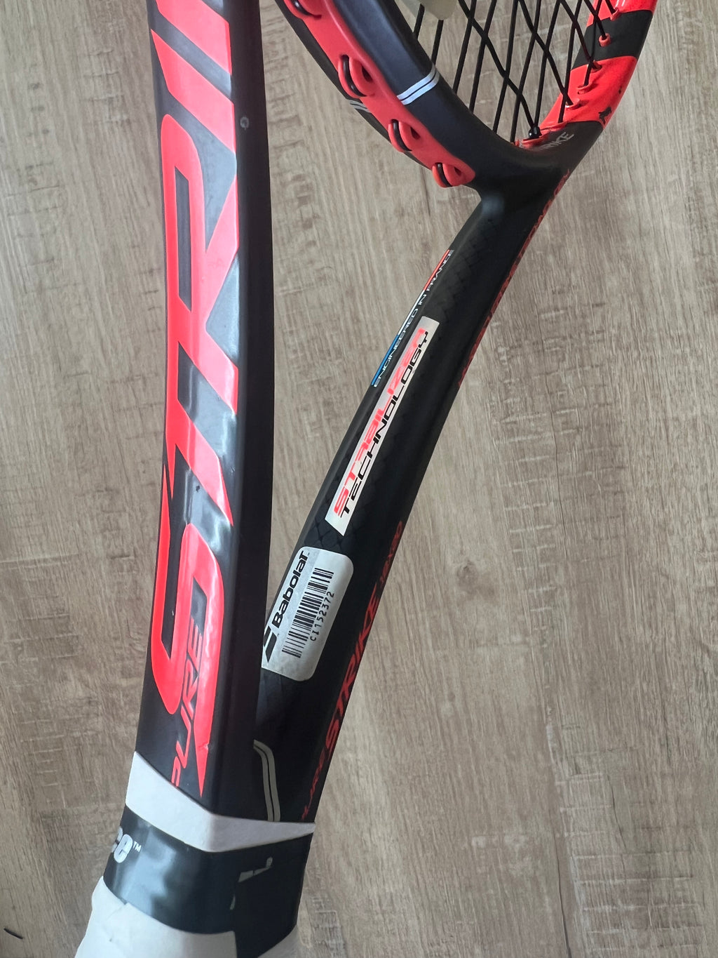 A close-up of a black and red Babolat tennis racket with "Pure Strike" and "Technifibre" branding, resting on a wooden surface.