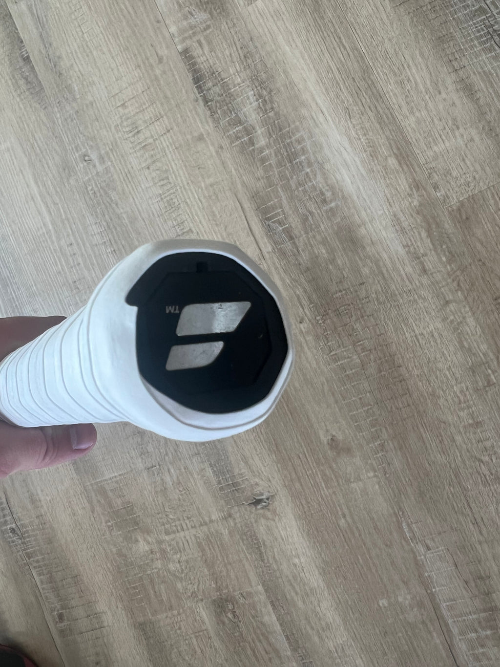 A hand holding a tennis racquet with a white grip, showing the black Babolat logo on the racquet’s butt cap against a wooden floor background.
