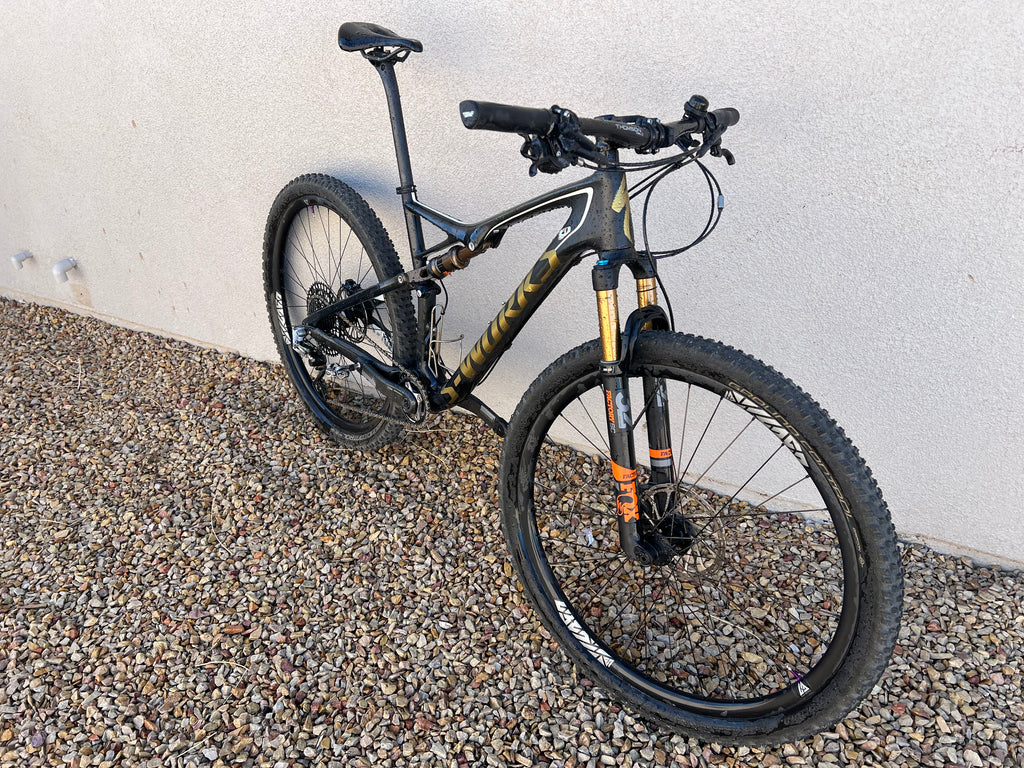 A Specialized S-WORKS Epic - Large, a black full-suspension mountain bike with gold accents, Fox Factory suspension, and LightCarbon wheels, is parked on gravel against a light-colored wall.