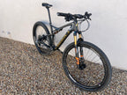 A Specialized S-WORKS Epic - Large, a black full-suspension mountain bike with gold accents, Fox Factory suspension, and LightCarbon wheels, is parked on gravel against a light-colored wall.