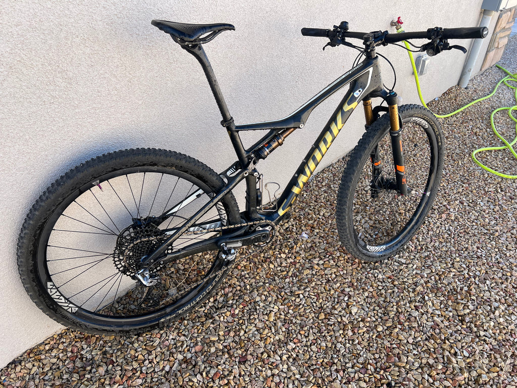 A black Specialized S-WORKS Epic - Large full-suspension mountain bike with yellow branding, Fox Factory suspension, and LightCarbon wheels is parked on gravel beside a beige wall and green garden hose.