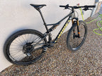 A black Specialized S-WORKS Epic - Large full-suspension mountain bike with yellow branding, Fox Factory suspension, and LightCarbon wheels is parked on gravel beside a beige wall and green garden hose.