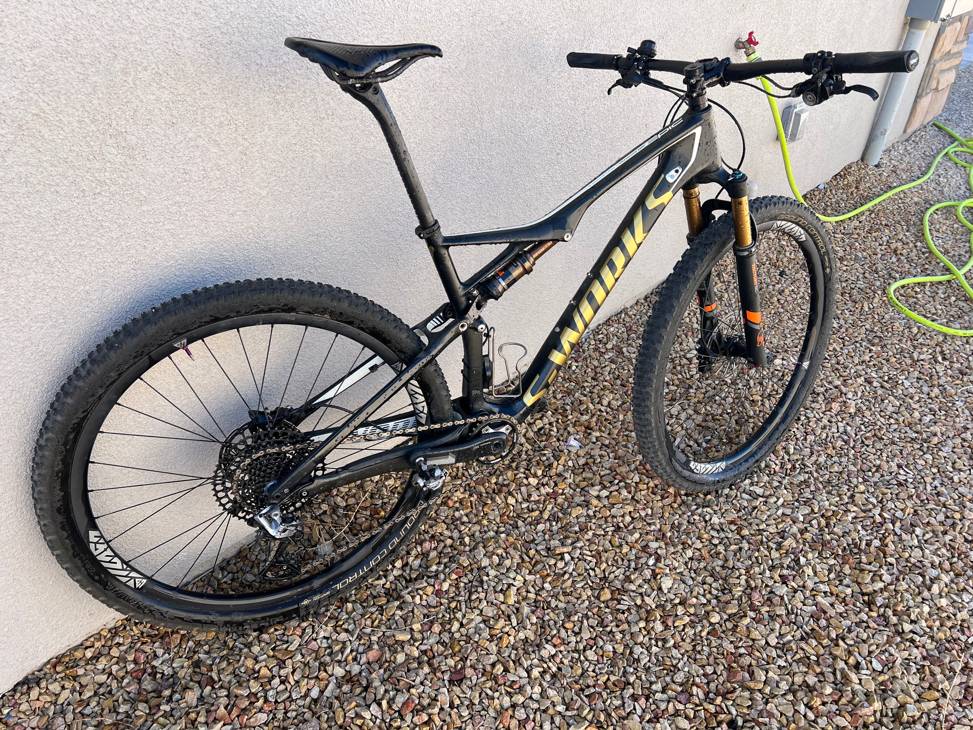A black Specialized S-WORKS Epic - Large full-suspension mountain bike with yellow branding, Fox Factory suspension, and LightCarbon wheels is parked on gravel beside a beige wall and green garden hose.