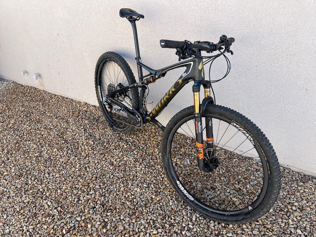 A Specialized S-WORKS Epic - Large full-suspension mountain bike in black and gold with Fox Factory suspension and LightCarbon wheels is parked on gravel by a light-colored wall.