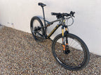A Specialized S-WORKS Epic - Large full-suspension mountain bike in black and gold with Fox Factory suspension and LightCarbon wheels is parked on gravel by a light-colored wall.