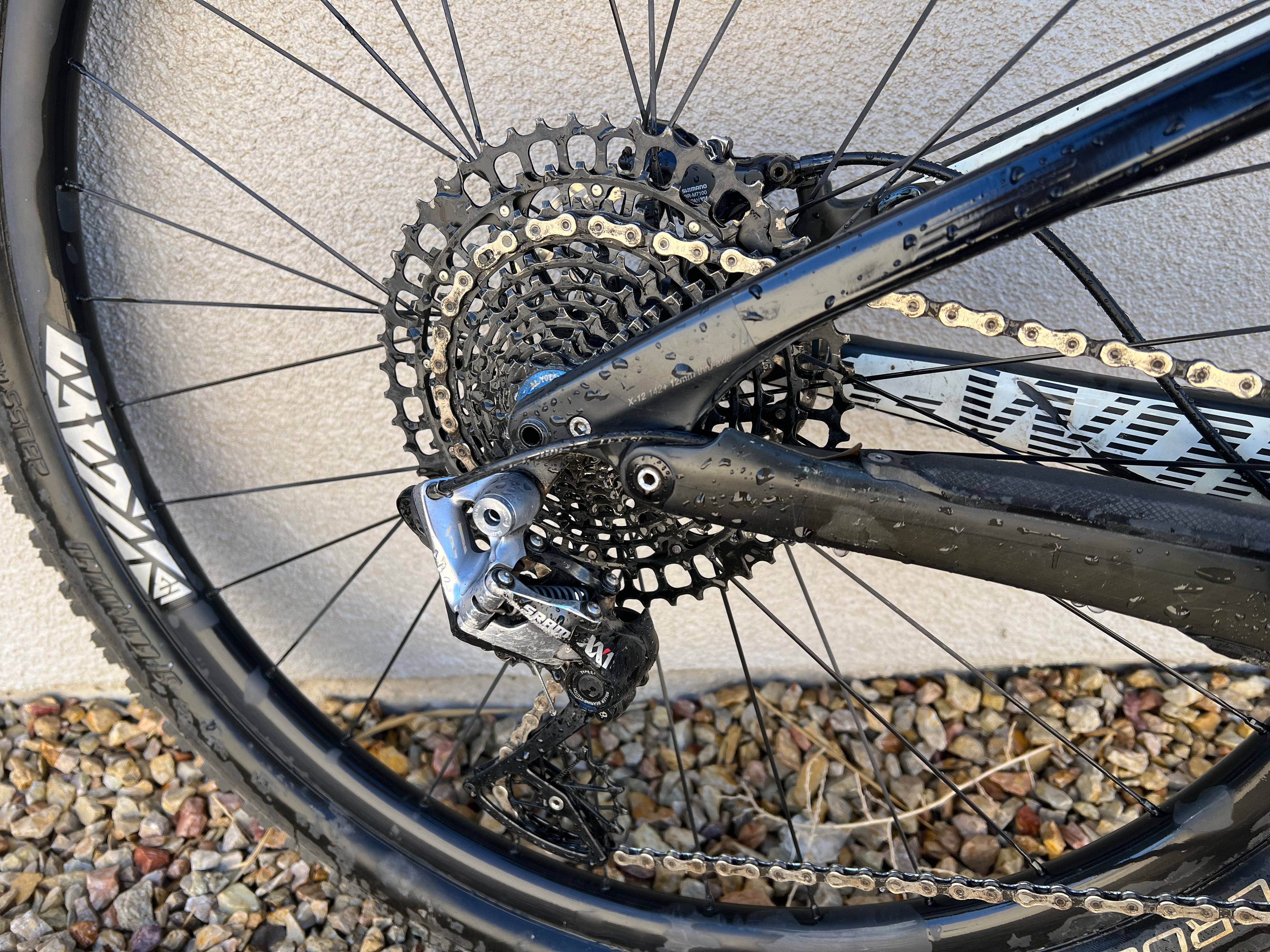 Close-up of a Specialized S-WORKS Epic (Large) rear wheel, showcasing gears, derailleur, and chain with water droplets on the frame and gravel scattered on the ground.